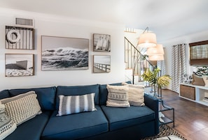A modern living room with a dark blue sofa, multiple framed artworks, a staircase with a unique railing, and a large floor lamp.