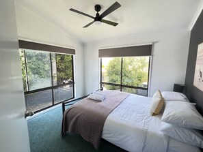 Une chambre à coucher avec un lit, un ventilateur de plafond, de grandes fenêtres et une vue sur les arbres.