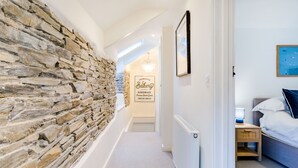 A hallway with a stone wall, a sign reading "Farmhouse Bakery Handmade Premium Baked Goods Fresh Daily," a bed with a blue headboard, and a nightstand with a lamp. - The Old Bakehouse - Within the Helland Barton Farm Collection (Delabole)