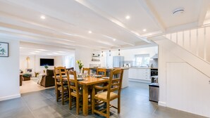 A spacious kitchen with a dining area, featuring a wooden table and chairs, a built-in oven, and a large island with a sink. - The Old Bakehouse - Within the Helland Barton Farm Collection (Delabole)