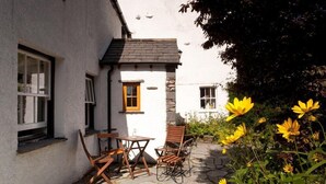 A white building with a stone facade, a small outdoor seating area with wooden chairs and tables, and yellow flowers in the foreground.