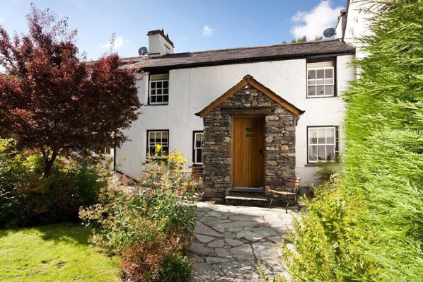 A traditional stone and plaster house with a stone entrance, a grassy garden, and a clear blue sky. - Bobbin Beck Cottage (Ulverston)
