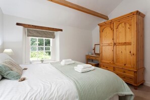 A bedroom with a large bed, a wooden wardrobe, a window with a view of greenery, and a lamp on the nightstand. - Bobbin Beck Cottage (Ulverston)