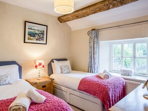 A bedroom with two beds, a wooden nightstand with a lamp, a window with curtains, and a framed picture on the wall.