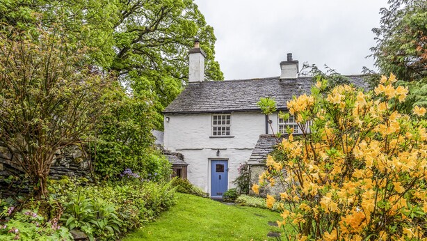 Ein traditionelles weißes Cottage mit blauer Tür, umgeben von üppigem Grün und blühenden Pflanzen.