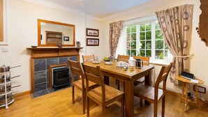 A dining room with a wooden table, chairs, a fireplace, and a window with curtains. - Uldale (Ambleside)