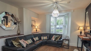 A modern living room with a black leather sectional sofa, a white armchair, a wooden floor, and a large window with shutters. - Newby Bridge House (Ulverston)