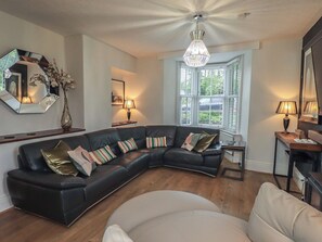 A modern living room with a black leather sectional sofa, a white armchair, a wooden floor, and a large window with shutters. - Newby Bridge House (Ulverston)