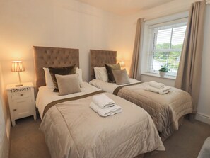 A hotel room with two beds, a window with curtains, a bedside table with a lamp, and a potted plant on the windowsill. - Newby Bridge House (Ulverston)