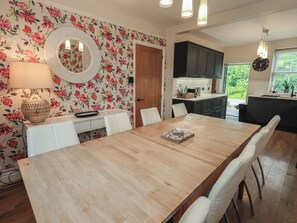 A dining area with a wooden table, white chairs, a floral wallpaper, and a kitchen in the background. - Newby Bridge House (Ulverston)