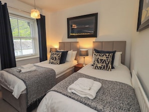 Quarto de hotel com duas camas, janela com cortinas, quadro na parede e uma luminária em cada mesa de cabeceira.