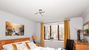 A hotel room with a bed, a desk, a chair, a window with curtains, a lamp, and a painting on the wall. - Spring Cottage (Ambleside)