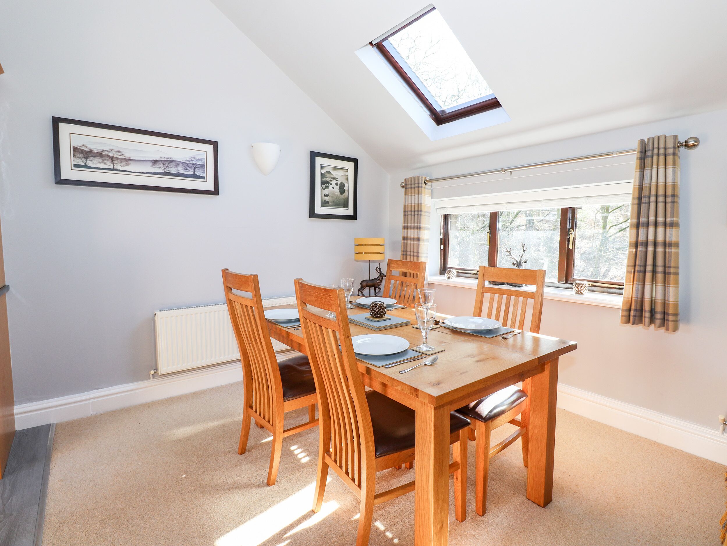 A dining area with a wooden table, four chairs, a skylight, a window with curtains, and framed pictures on the wall.