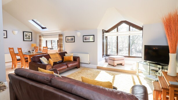 A spacious living room with a high ceiling, a large window, a brown leather sofa, a dining area with wooden chairs, and a television on a glass table. - Spring Cottage (Ambleside)