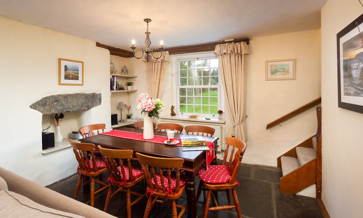 A dining area with a wooden table, chairs, and a chandelier. There is a window with curtains and a view of greenery outside.