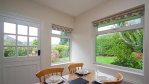 A dining room with a wooden table set for four, large windows with curtains, and a view of a garden.