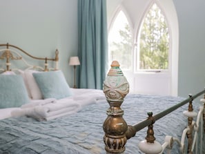 Una cama con colcha azul con textura, cabecero metálico decorativo y ventana con vistas a árboles.