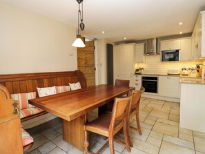 Un comedor con una mesa y sillas de madera, un banco con cojines, una lámpara colgante y una cocina al fondo.