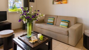 Una sala de estar con un sofá color beige, una mesa de centro con libros y un florero, un televisor y un ventanal con vista a árboles.