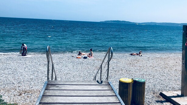 A wooden jetty leading to a pebble beach with a view of the sea and distant hills.