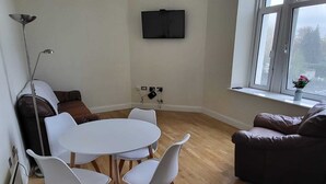 A room with a round table, four white chairs, a brown sofa, a floor lamp, and a TV mounted on the wall. - Stunning 3-bed Apartment in Heart of Cardiff Bay (Cardiff)
