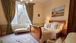 A bedroom with a wooden bed, a bedside table, a lamp, a window with curtains, a painting on the wall, and a chair.
