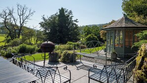 A gazebo with a green roof and white trim, surrounded by a garden with benches and a barbecue grill.