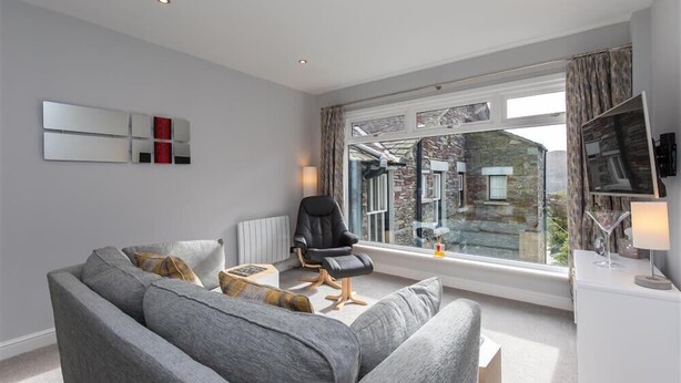 A modern living room with a grey sofa, a black chair, and a large window offering a view of a stone building. - Silver Howe (Ambleside)