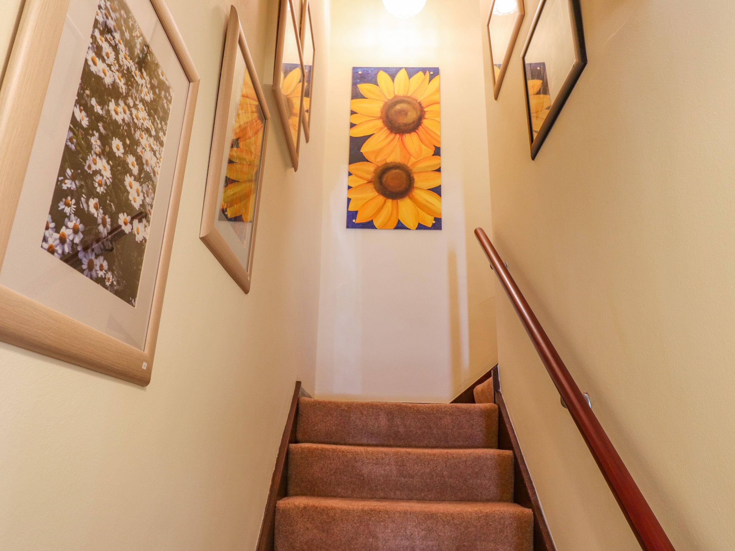 Un escalier avec une rampe en bois, des marches recouvertes de moquette et des tableaux représentant des motifs floraux et des tournesols accrochés aux murs.