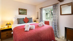 Una cama bien tendida con una colcha roja, dos almohadas y toallas enrolladas sobre la cama. Hay una mesita de noche con una lámpara y un cajón pequeño. Se ve una ventana con cortinas estampadas, y un cuadro cuelga en la pared sobre la cama.