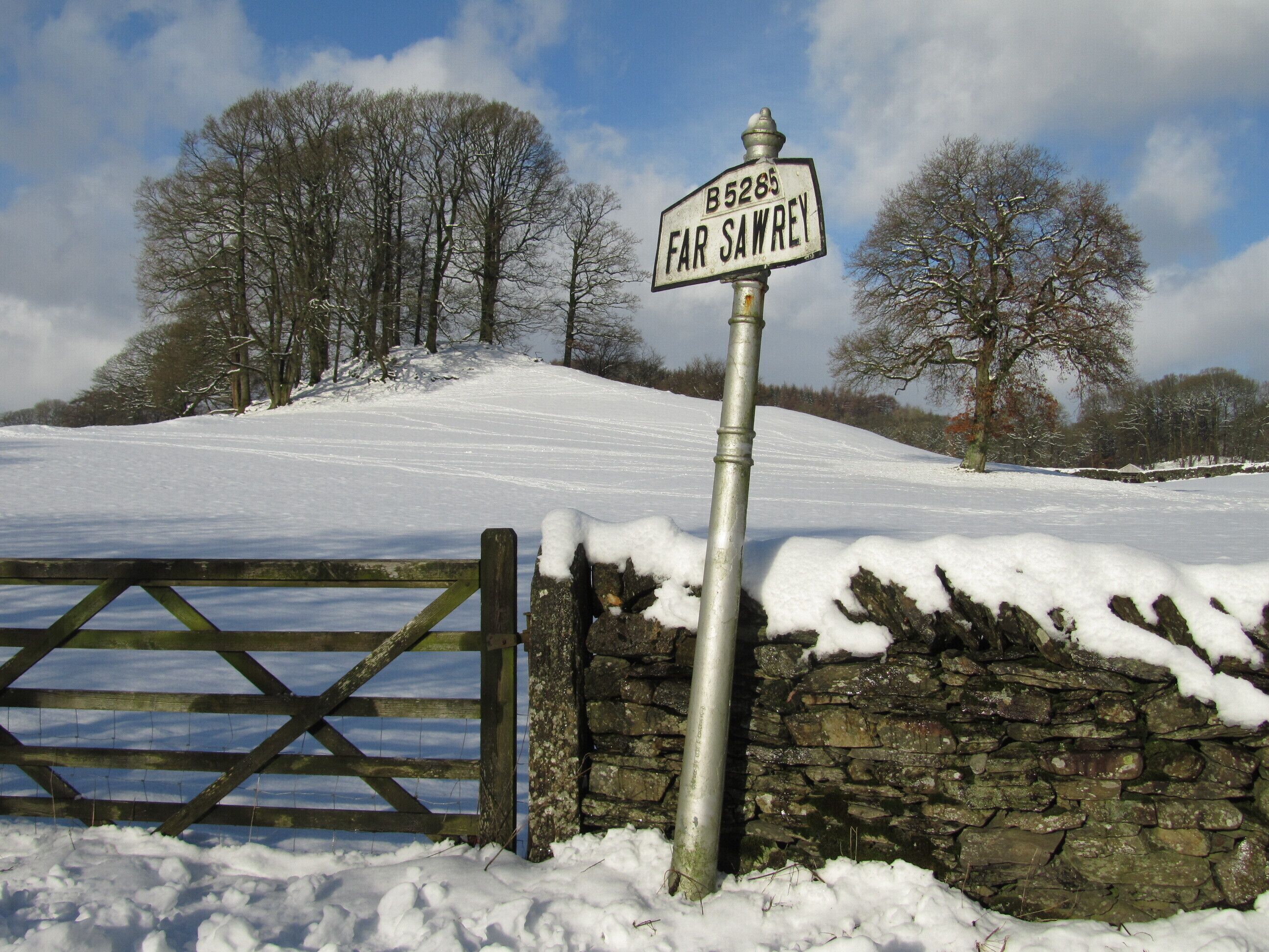 A rural landscape with a stone wall and a metal gate, a signpost indicating 'B5285 Far Sawrey', and a snow-covered field with trees in the background.