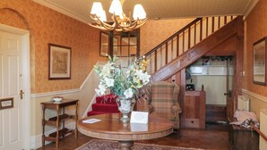 A grand entryway with a wooden staircase, a round wooden table with a floral centerpiece, and a red carpet leading to a door. - The Rockery Suite (Ambleside)