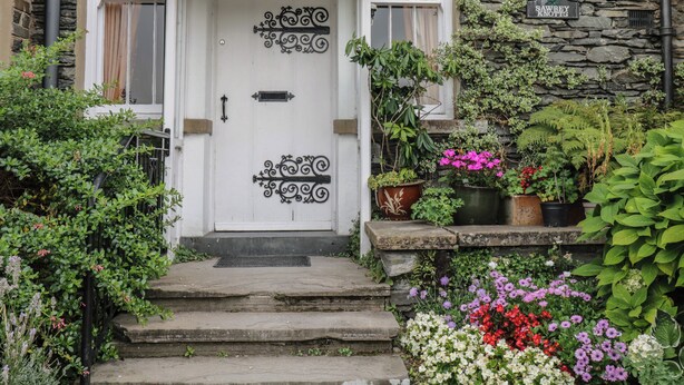 A white door with black ironwork, flanked by windows with curtains, leading to a stone building with a sign reading "SAWREY KNOTT'S". - The Rockery Suite (Ambleside)