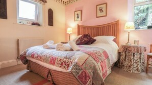 A bedroom with a large bed, a window with a floral curtain, a wooden headboard, and a bedside table with a lamp. - The Rockery Suite (Ambleside)