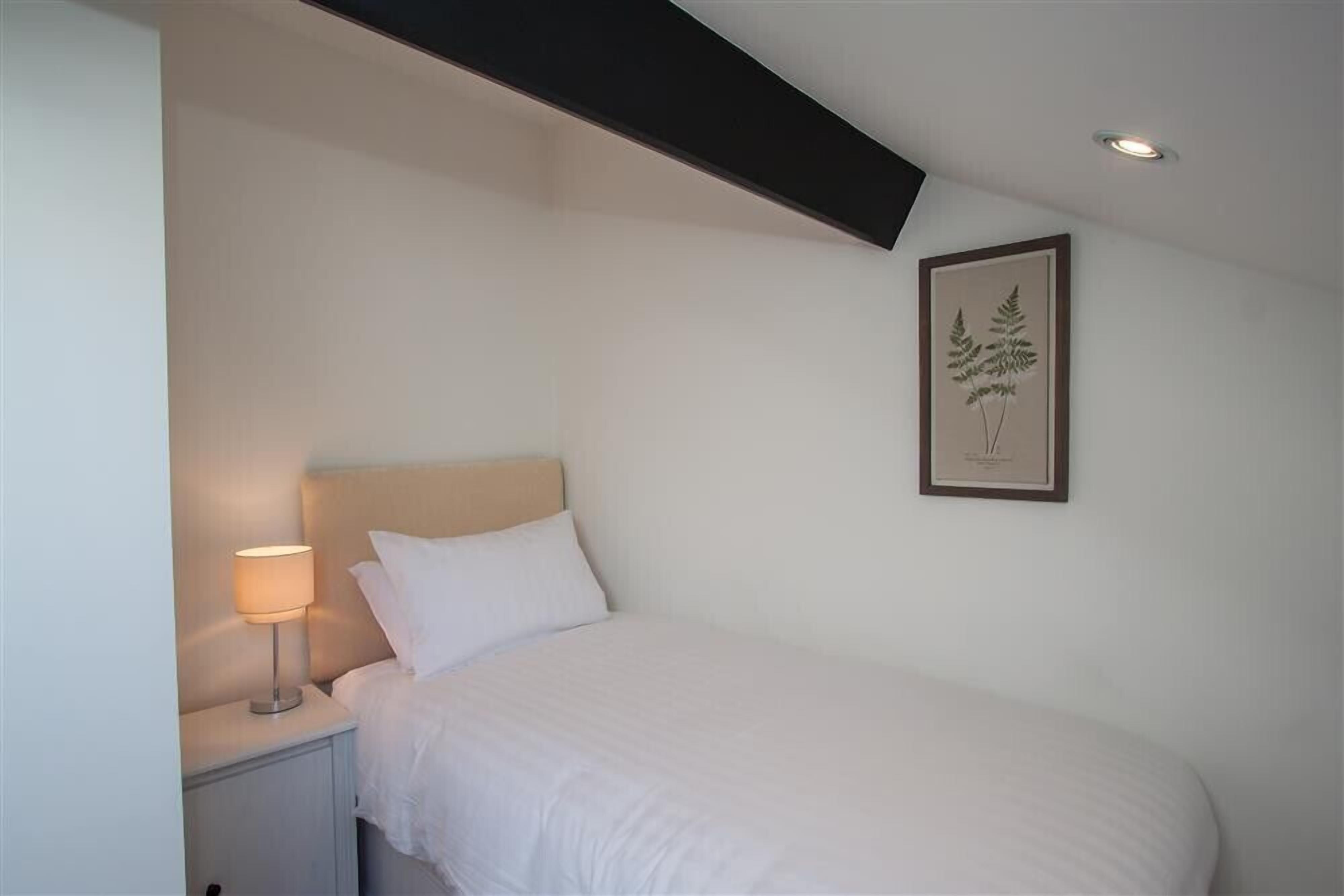 Ein Schlafzimmer mit Bett, Nachttisch mit Lampe, einer gerahmten Pflanze an der Wand und einer Deckenleuchte.