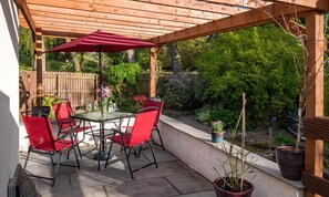 Un patio al aire libre con una mesa de vidrio y sillas rojas, rodeado por una pérgola de madera y plantas en macetas.