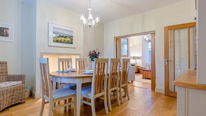 Un coin repas avec une table ronde, six chaises et un lustre. On aperçoit, à l’arrière-plan, une chaise en osier et une petite table avec une lampe.