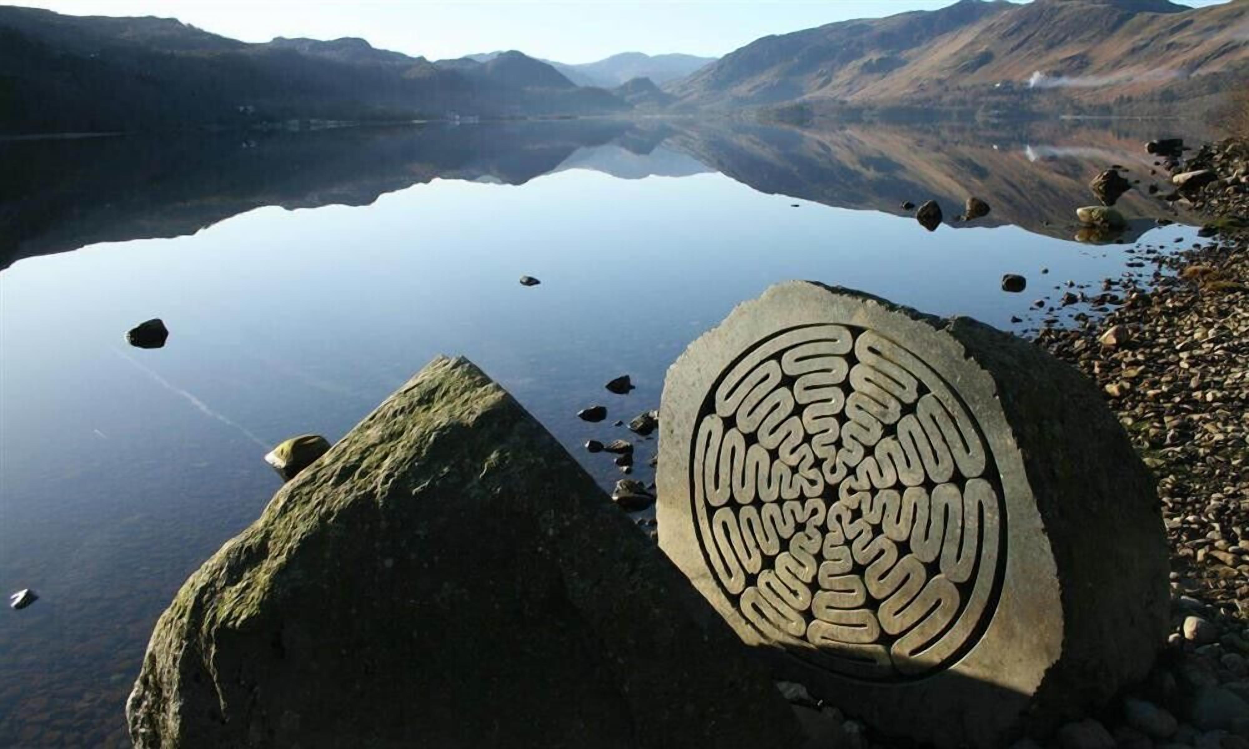 A large rock with a spiral pattern near a calm lake surrounded by mountains.