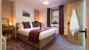 A neatly arranged bedroom with a bed, bedside tables, a nightstand, a window with curtains, and a door. - The Cats Bield (Keswick)