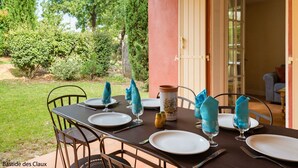 Un espace de restauration en plein air avec une table dressée pour quatre personnes, entourée de chaises, et une vue sur le jardin.