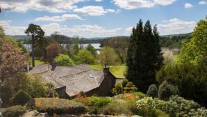 A traditional stone house with a sloped roof, surrounded by lush greenery and a view of a lake. - Betamere (Ambleside)