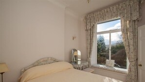 A bedroom with a large bed, a bedside table with a lamp, a dresser with a mirror, and a window with curtains. - Betamere (Ambleside)