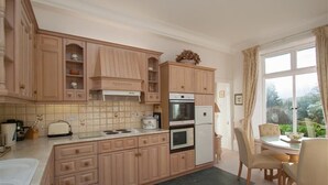 A kitchen with wooden cabinets, a stove, oven, and a refrigerator. There is a dining table with chairs and a window with curtains. - Betamere (Ambleside)