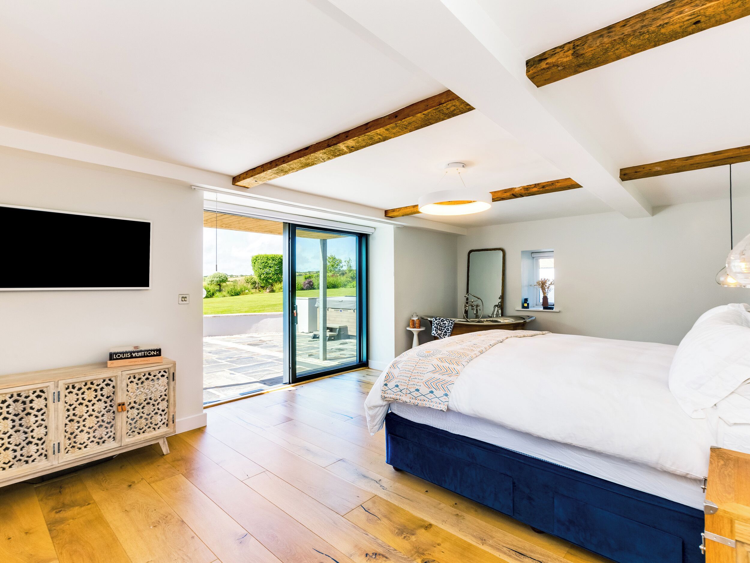 Ein modernes Schlafzimmer mit einem großen Bett, einem an der Wand montierten Fernseher, einer Schiebeglastür zu einem Außenbereich und einer Holzbalkendecke.
