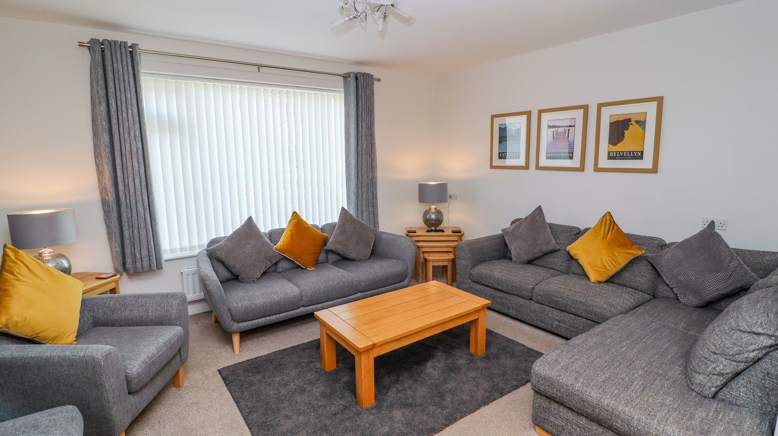 A modern living room with grey sofas, a wooden coffee table, and a large window with curtains. There are three framed pictures on the wall.
