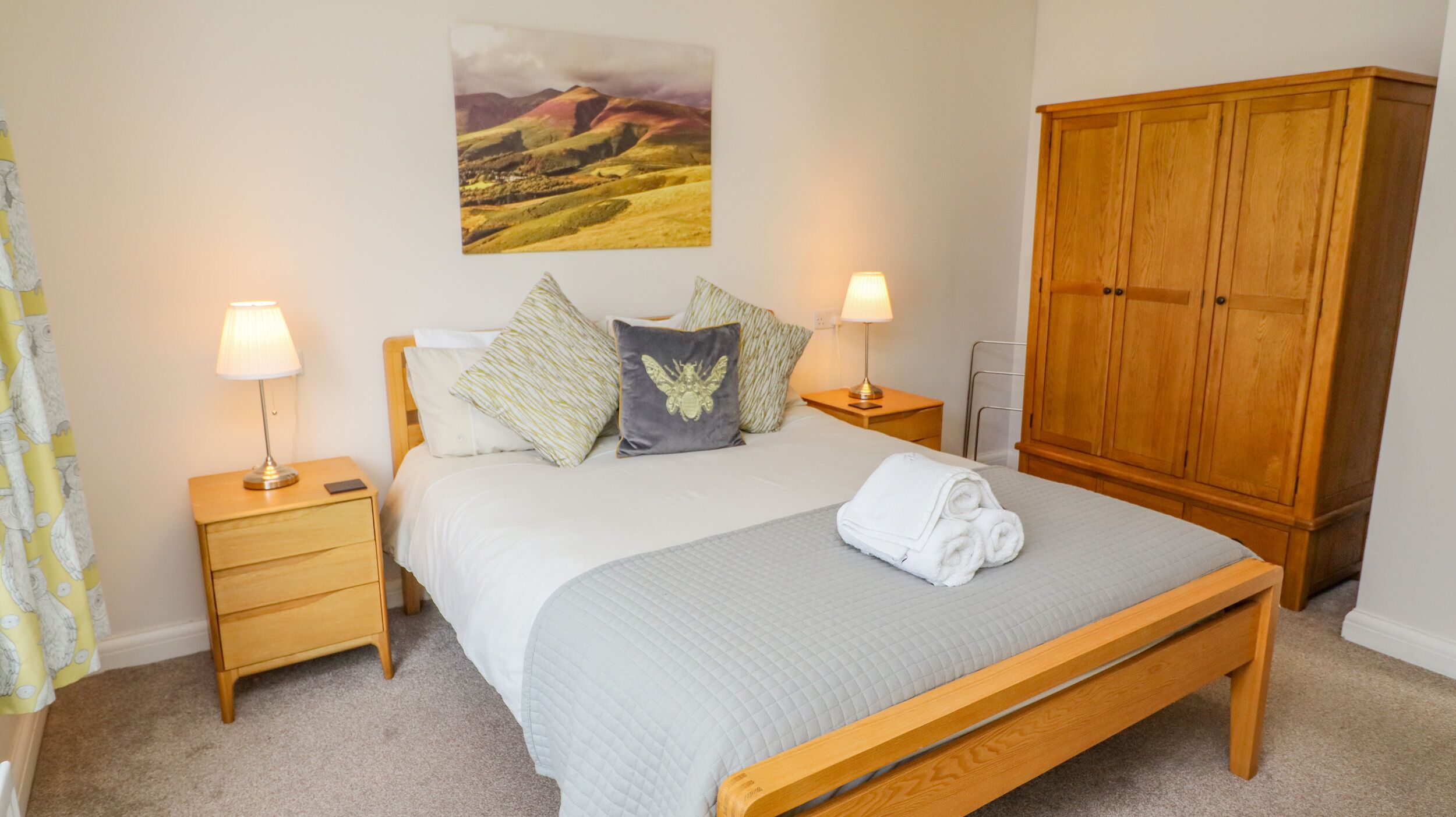A neatly made bed with a quilted grey bedspread, two bedside tables with lamps, a wooden wardrobe, and a landscape painting on the wall.