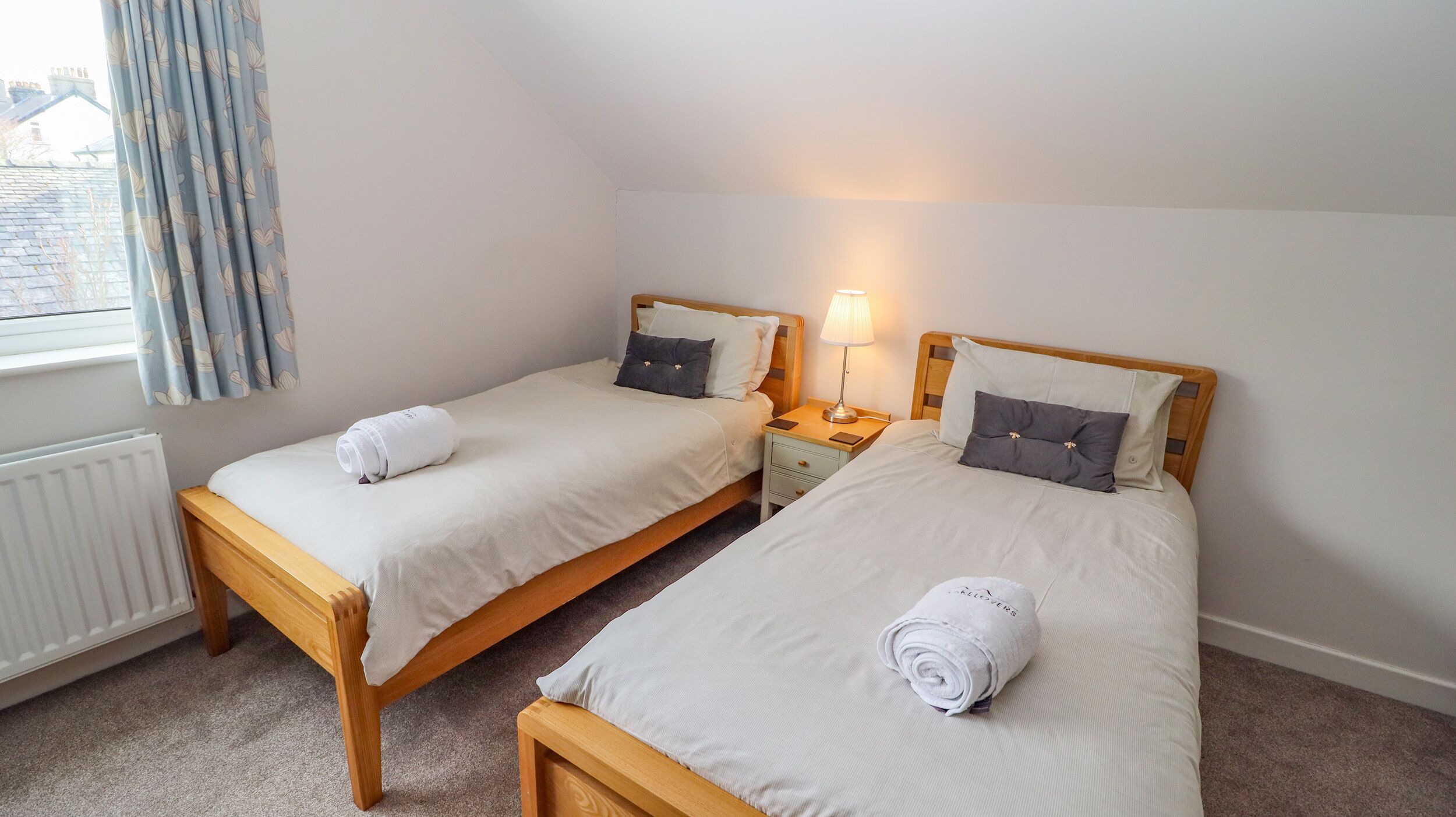 Une chambre d’hôtel avec deux lits simples, chacun doté d’une tête de lit en bois, d’une table de chevet avec une lampe et d’une serviette roulée sur chaque lit.