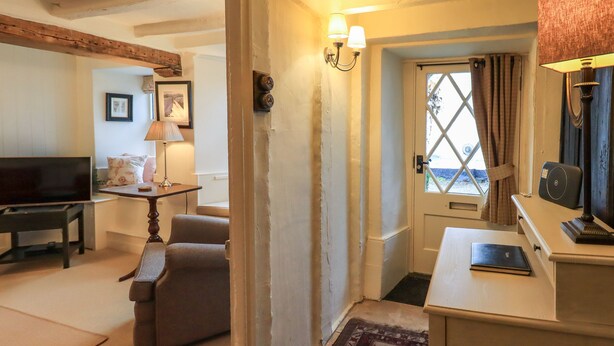 Una sala de estar con televisor, sofá y una pequeña mesa con lámpara. Hay una puerta que da a otra habitación y una alfombra en el suelo.