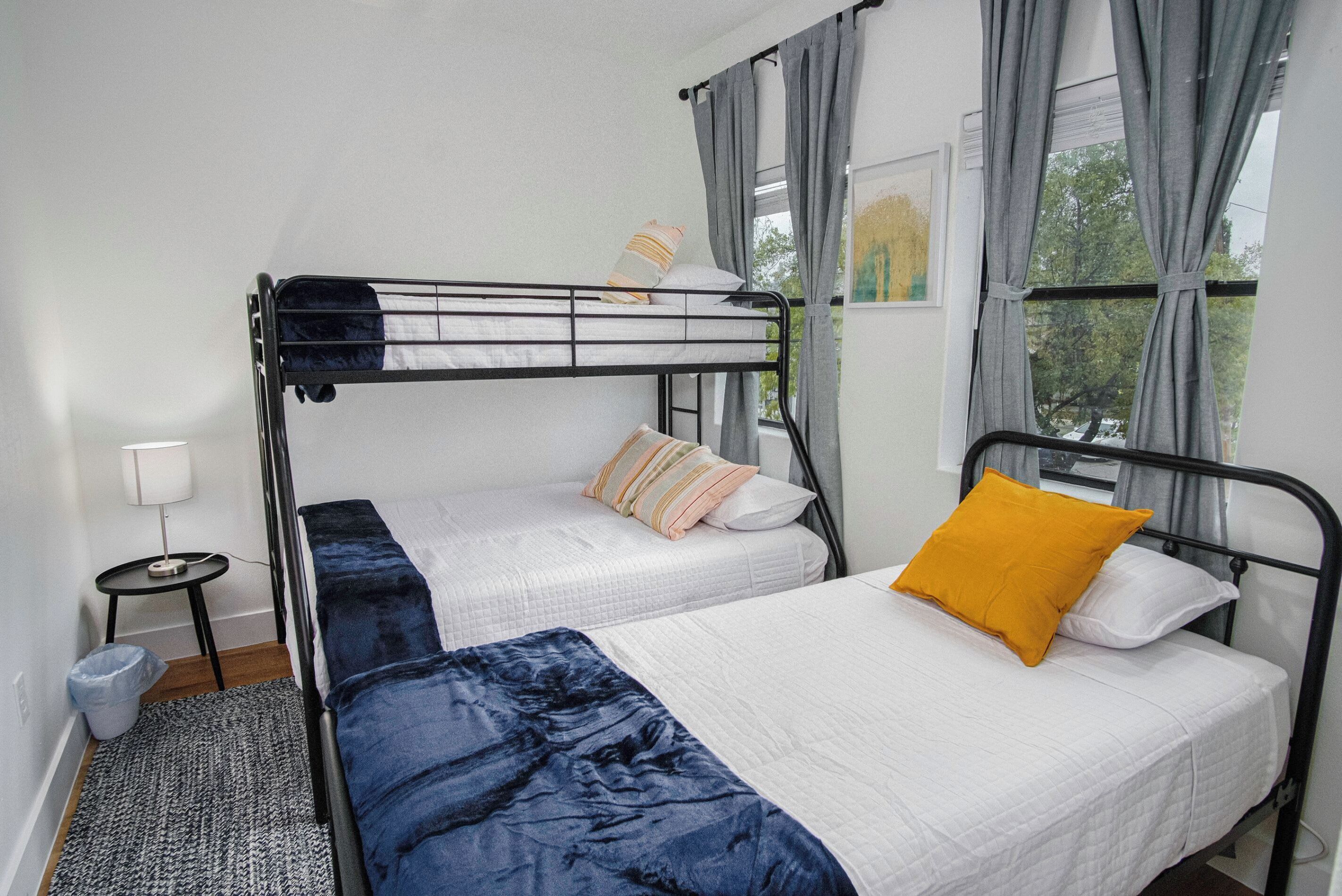 A dorm-style room with bunk beds, a small bedside table, a lamp, and a view of trees through the window.