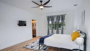 Une chambre à coucher comprenant un lit, un ventilateur de plafond, une télévision, une fenêtre avec des rideaux et une petite table avec une lampe.